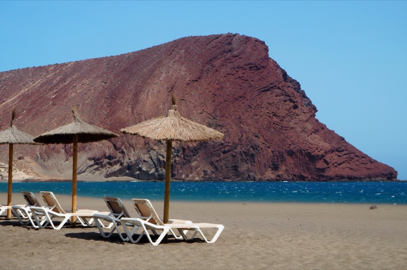 Red rock formation in the background of a sandy beach with straw umbrellas