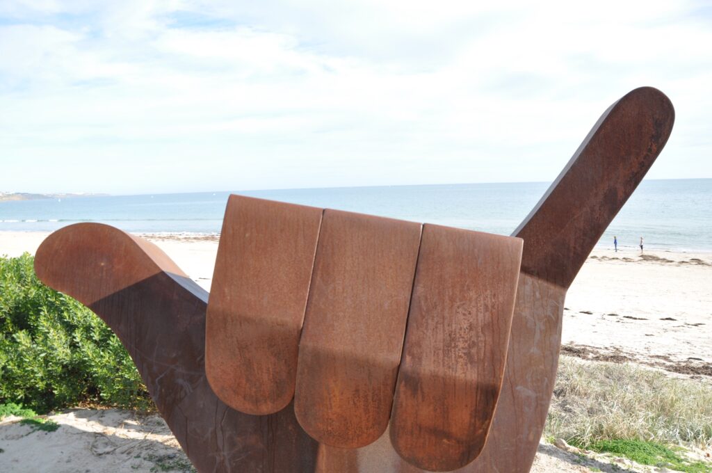 Giant metal sculpture of a hand making the 'Hang loose' sign in front of a beach in Adelaide, South Australia